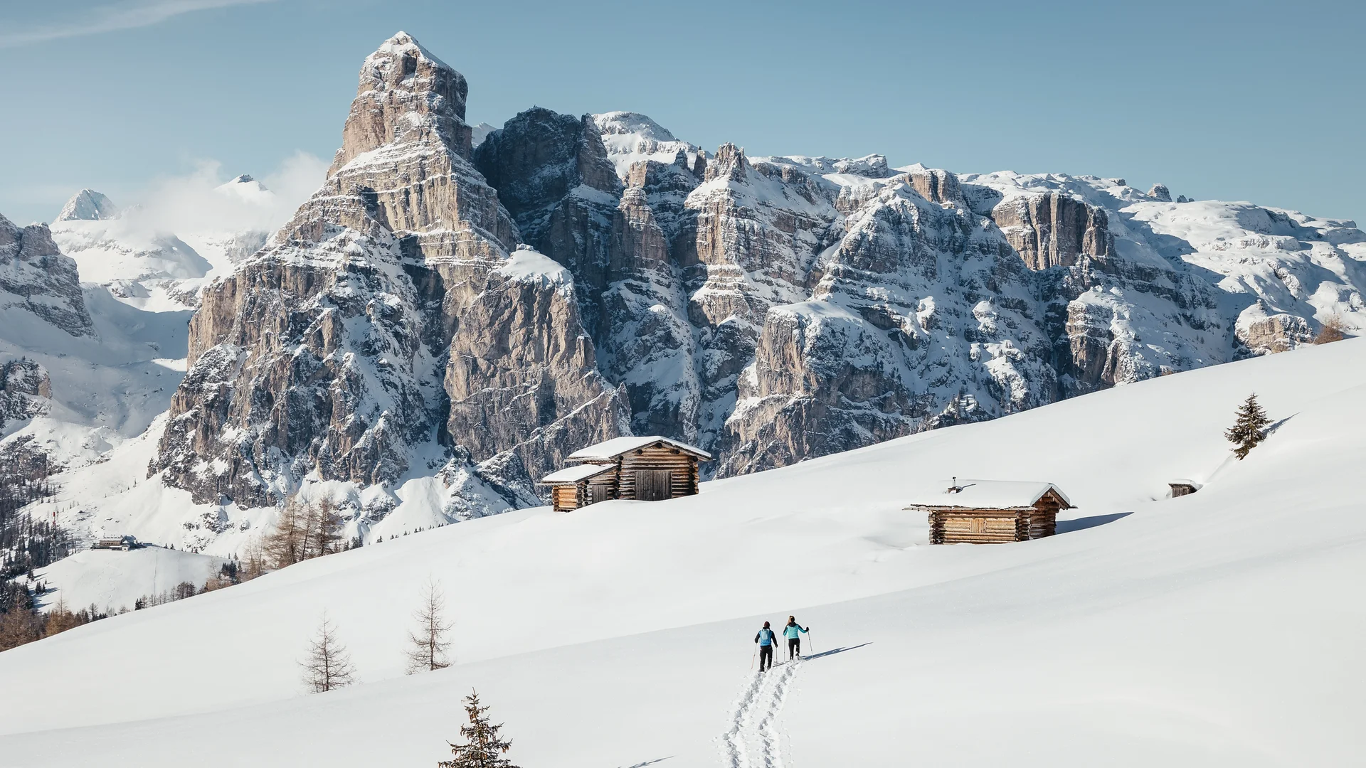 Zwei Skifahrer auf verschneiter Alm mit Holzhütten vor schneebedeckten Bergen