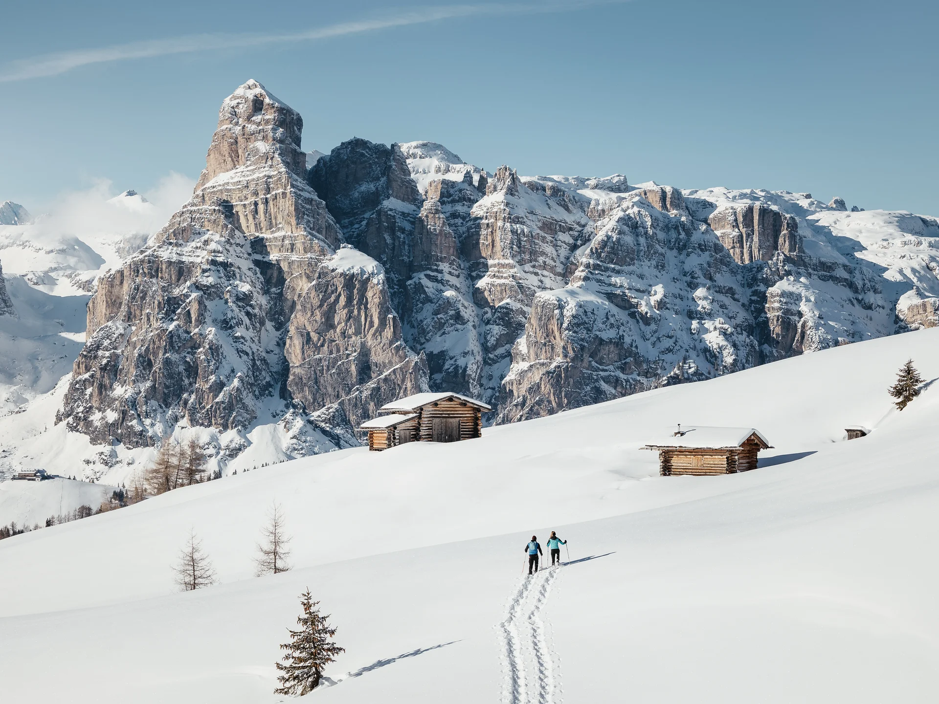 Zwei Skifahrer auf verschneiter Alm mit Holzhütten vor schneebedeckten Bergen