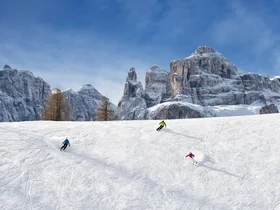 Den Winter in Alta Badia genießen – im Nature Hotel Delta Drei Skifahrer fahren auf schneebedeckter Piste vor felsigen Bergen