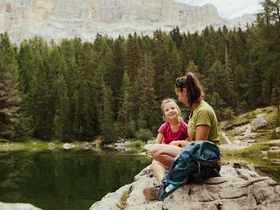 Auf zum Wandern in Alta Badia, rings ums Nature Hotel Delta! Mutter und Tochter sitzen auf Felsen am See vor Wald und Bergen