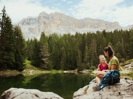 Mutter und Tochter sitzen auf Felsen am See vor Wald und Bergen