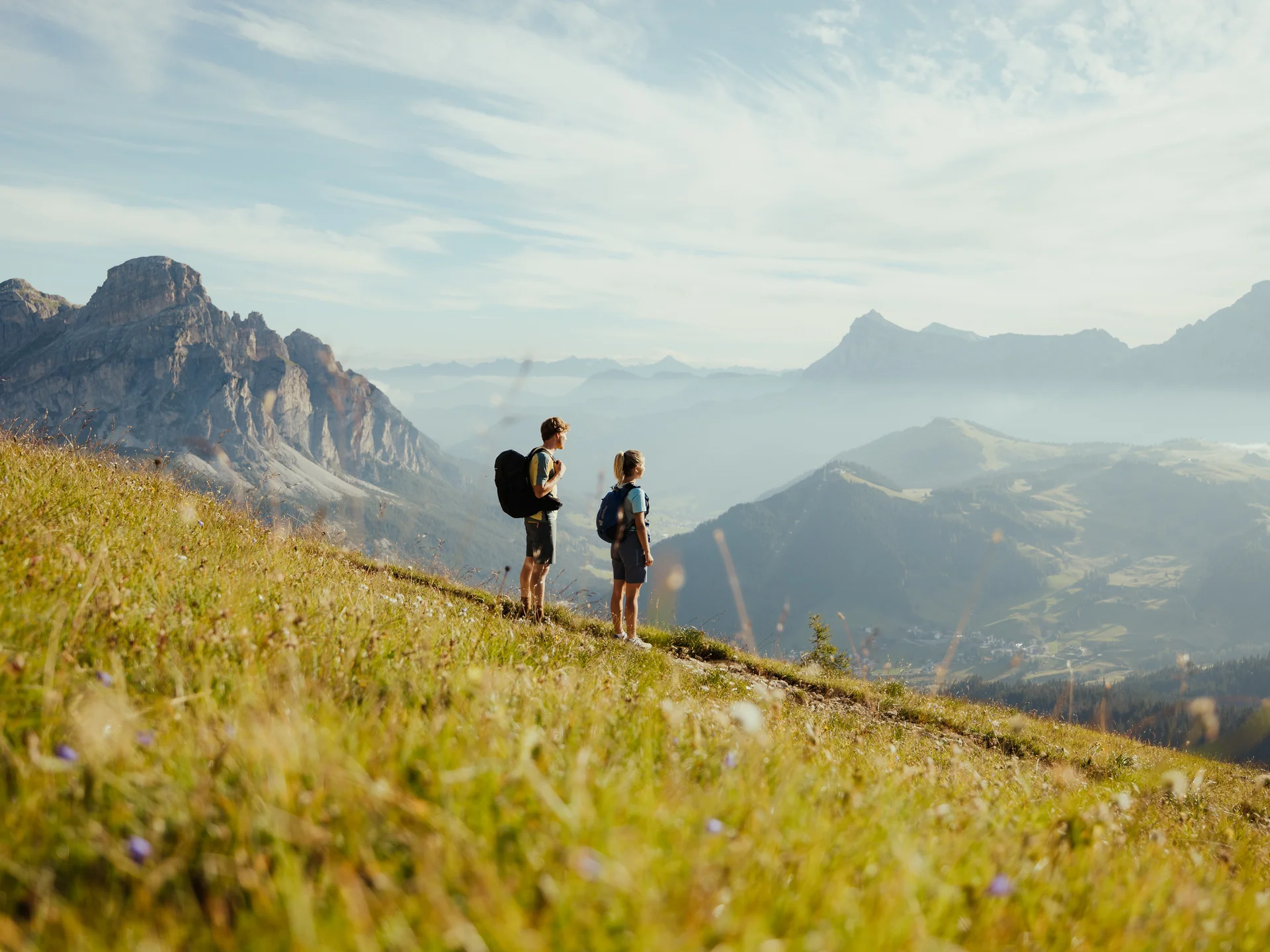 Zwei Wanderer mit Rucksäcken auf Bergwiese mit Blick auf Berge und Tal