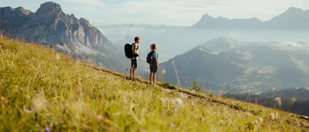Zwei Wanderer mit Rucksäcken auf Bergwiese mit Blick auf Berge und Tal