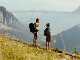 Auf zum Wandern in Alta Badia, rings ums Nature Hotel Delta! Zwei Wanderer mit Rucksäcken auf Bergwiese mit Blick auf Berge und Tal