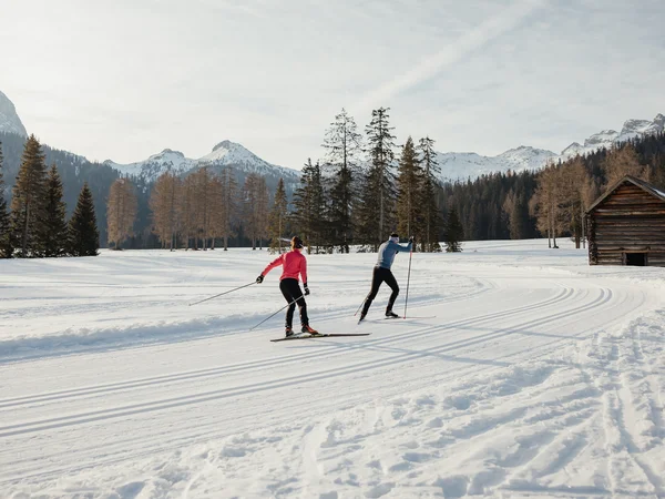 Zwei Langläufer auf verschneiter Loipe vor Bergkulisse und Holzhütte