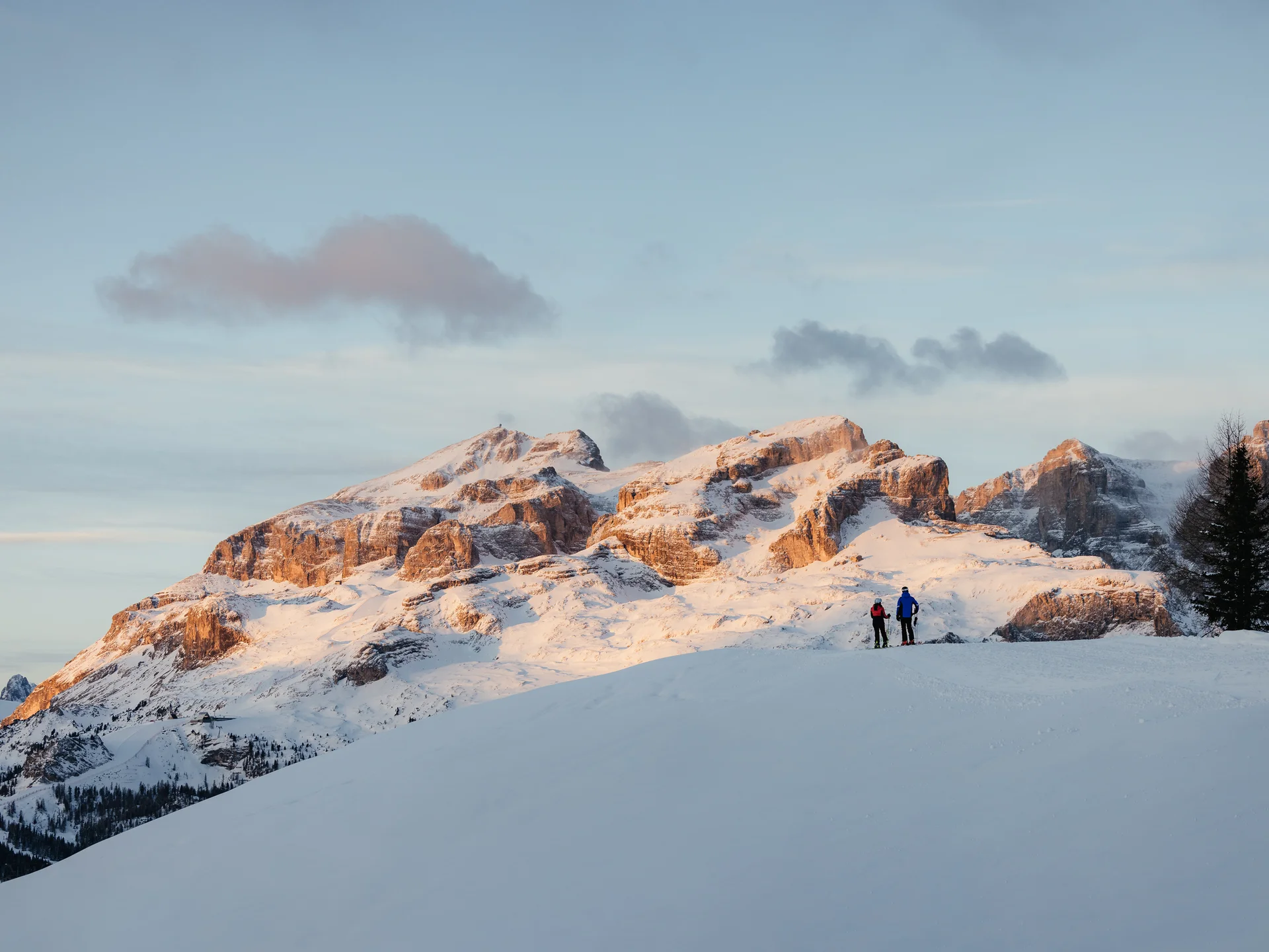 Zwei Skifahrer auf schneebedecktem Hügel mit Sonnenuntergang über Bergen