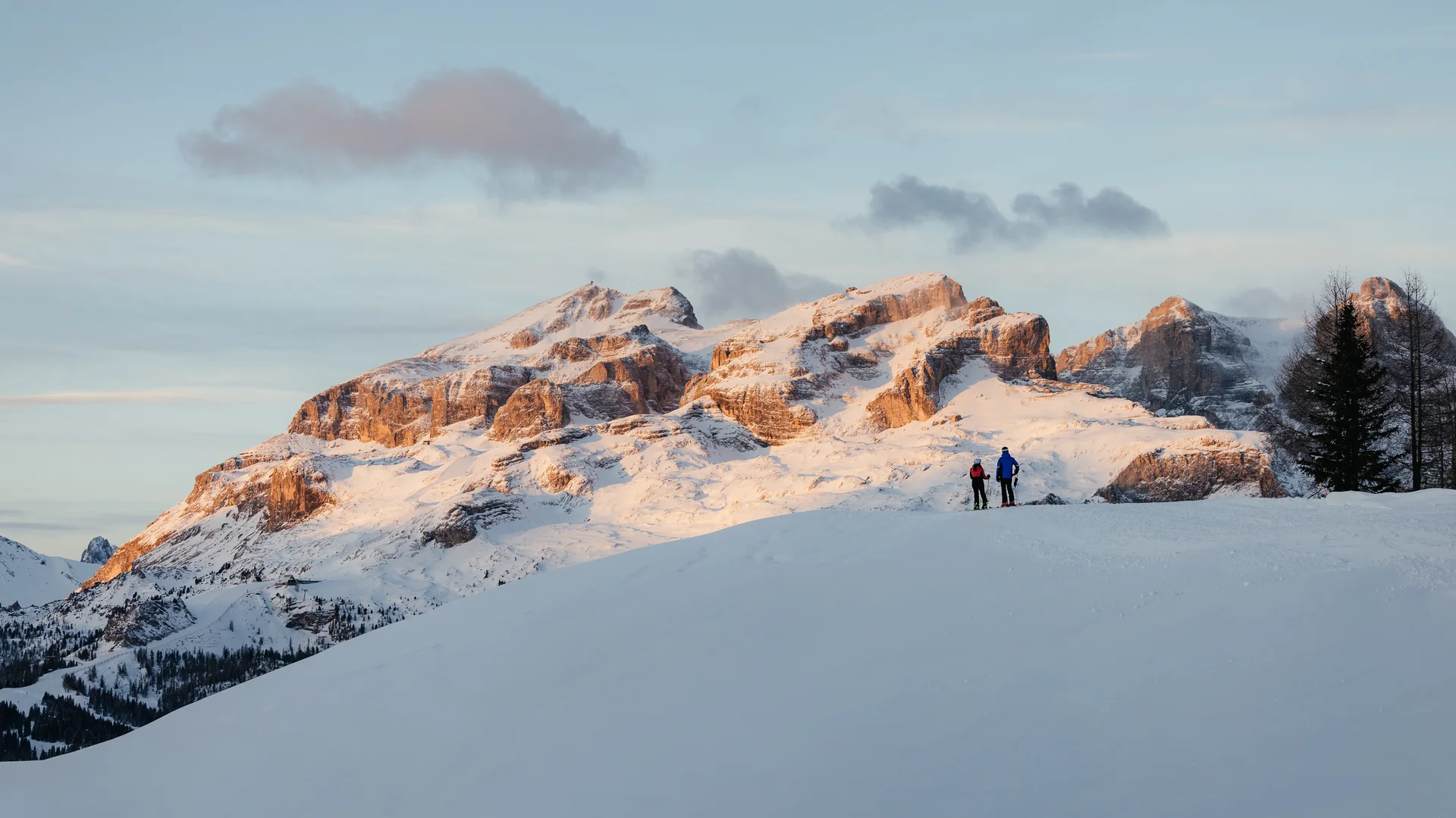 Den Winter in Alta Badia genießen – im Nature Hotel Delta Zwei Skifahrer auf schneebedecktem Hügel mit Sonnenuntergang über Bergen