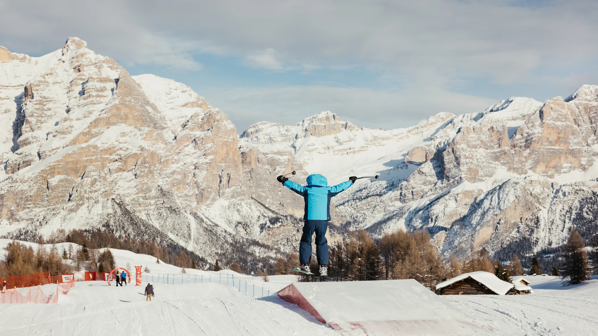 Die schönsten Eindrücke aus dem Nature Hotel Delta Kind in blauer Skibekleidung springt mit Skiern vor schneebedeckten Bergen