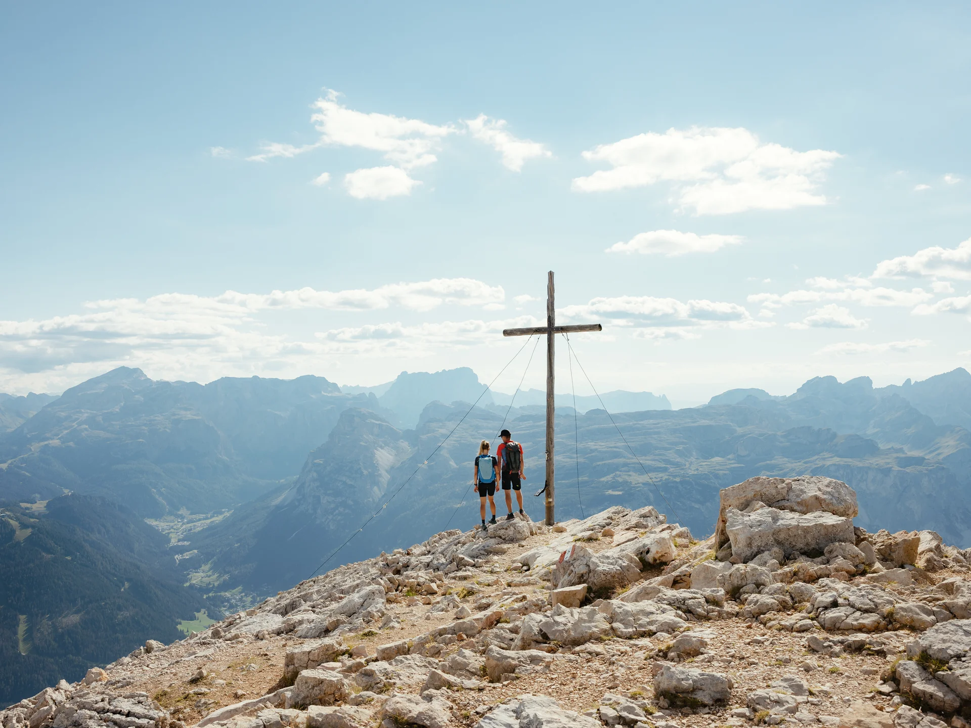 Zwei Wanderer stehen neben Gipfelkreuz mit Bergpanorama im Hintergrund