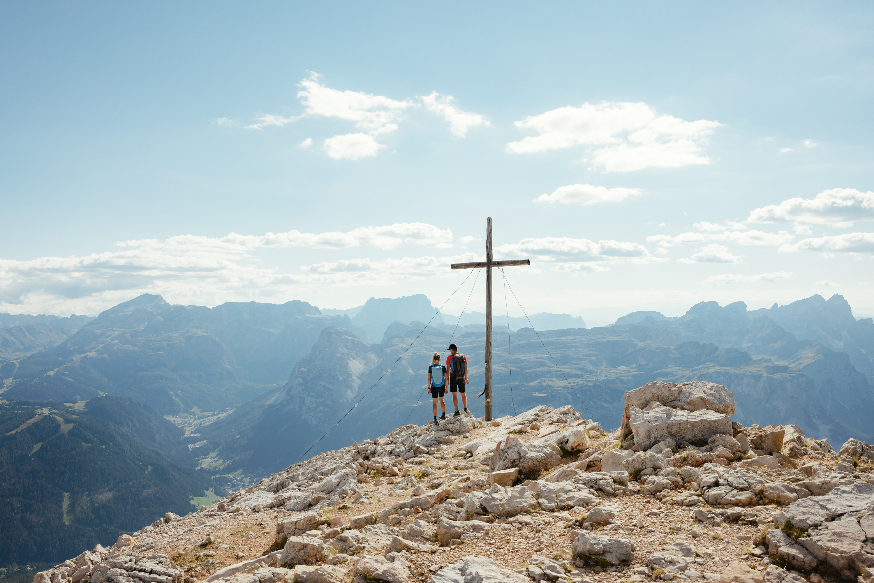 Auf zum Wandern in Alta Badia, rings ums Nature Hotel Delta! Zwei Wanderer stehen neben Gipfelkreuz mit Bergpanorama im Hintergrund