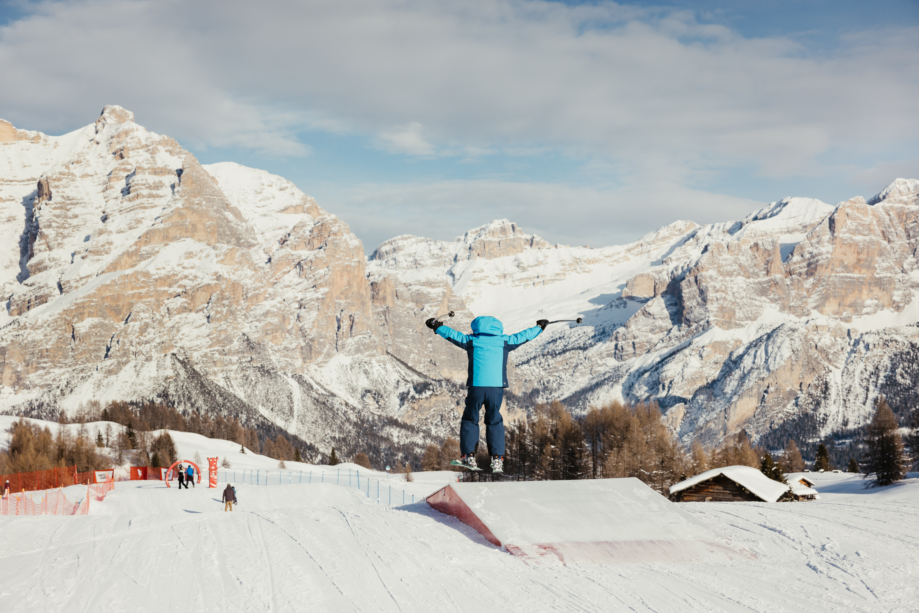 Le vostre vacanze in Alta Badia al Nature Hotel Delta Bambino con abbigliamento da sci blu salta sugli sci davanti a montagne innevate